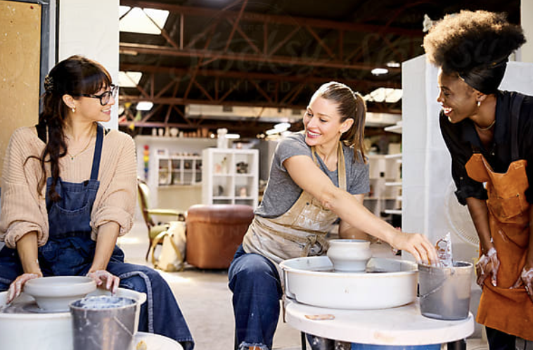 Three women laughing together around pottery wheels in a ceramics studio